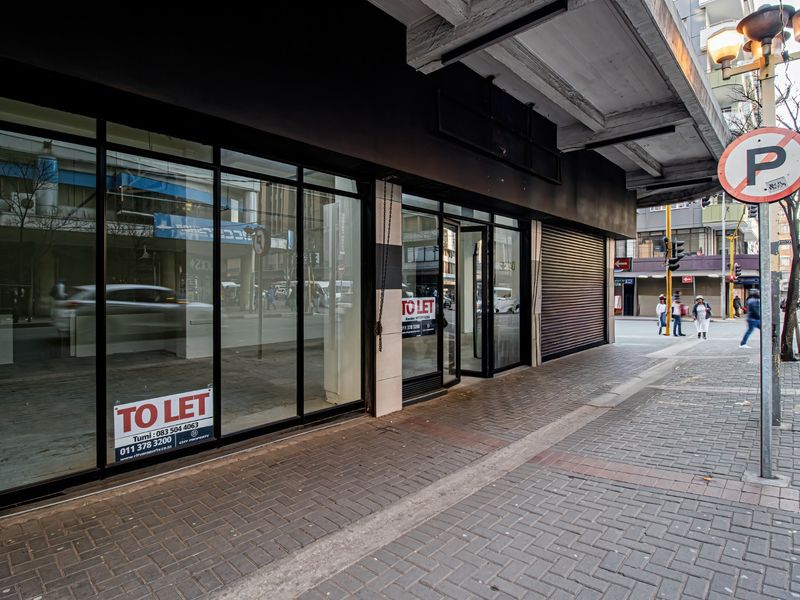 Commercial storefront with large glass display windows, metal roller shutter, and paved sidewalk access in a busy urban street setting.