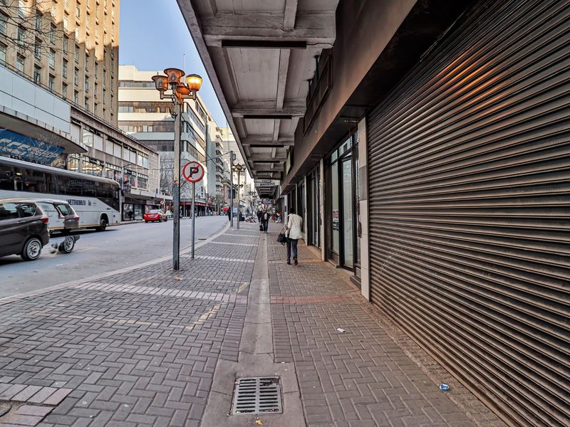 A street-level view of a commercial building exterior featuring a paved sidewalk, metal roller shutters, and urban street infrastructure.