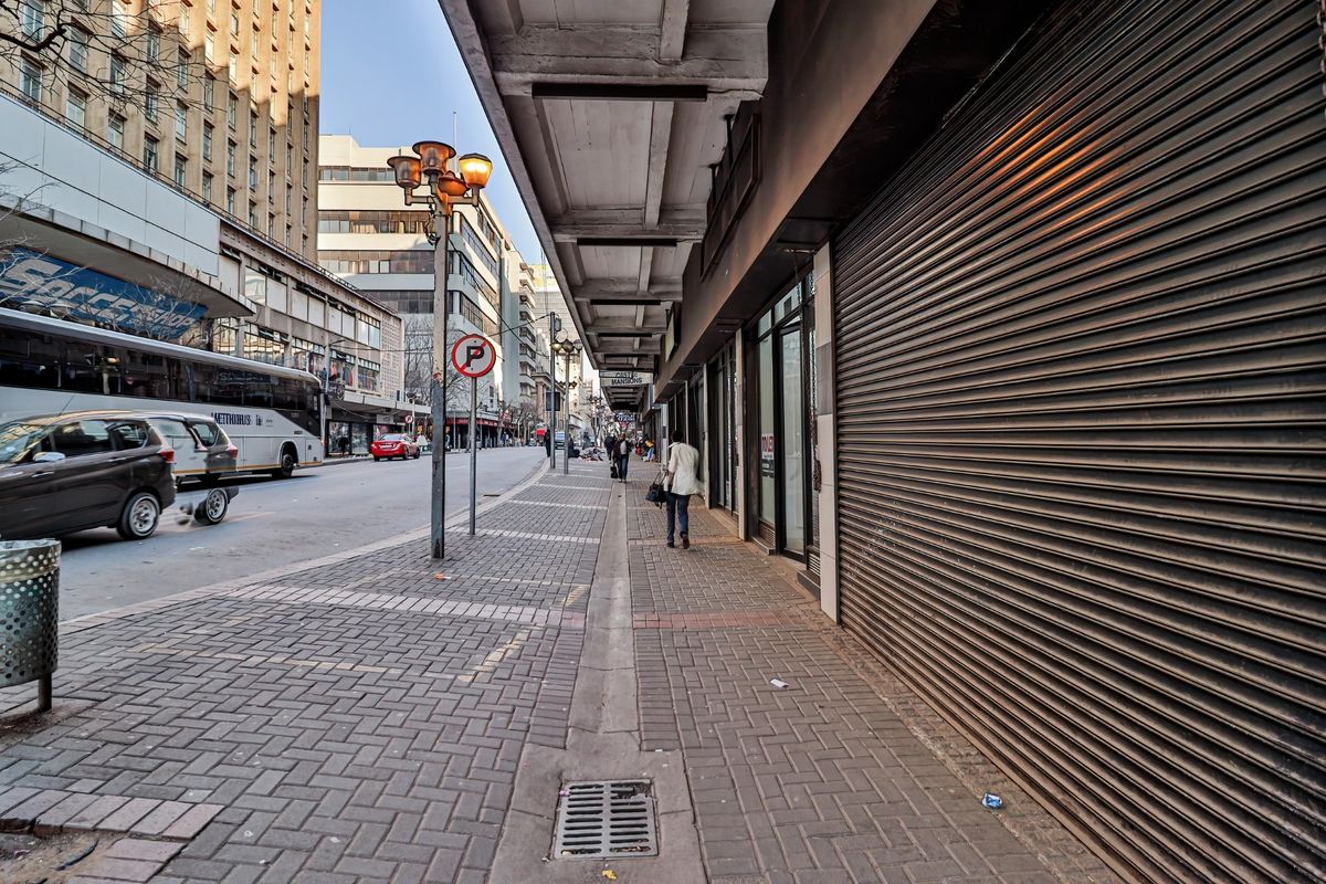 A street-level view of a commercial building exterior featuring a paved sidewalk, metal roller shutters, and urban street infrastructure.