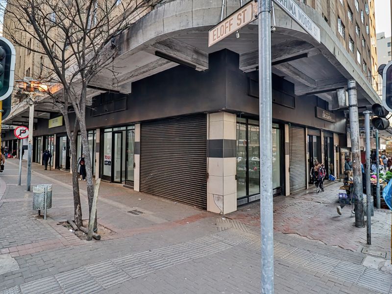 Corner commercial retail space featuring large display windows and metal roller shutters, located at a busy street intersection.