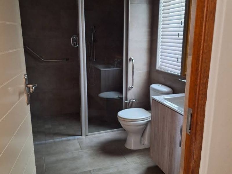 A functional bathroom featuring a walk-in shower with glass enclosure, grab rails, a vanity cabinet with basin, and neutral floor tiling.