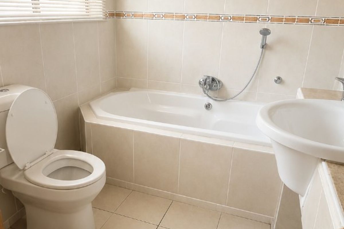 A functional bathroom featuring a built-in bathtub, pedestal washbasin, toilet, and neutral ceramic tiling throughout.