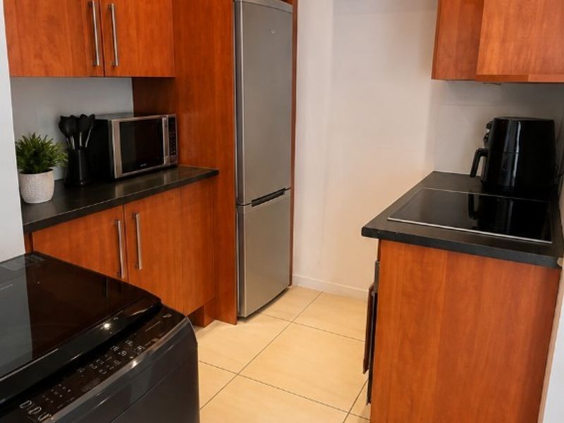 A compact kitchen featuring warm-toned wood cabinetry, dark countertops, ceramic floor tiles, and integrated appliances including an electric cooktop and extractor fan.