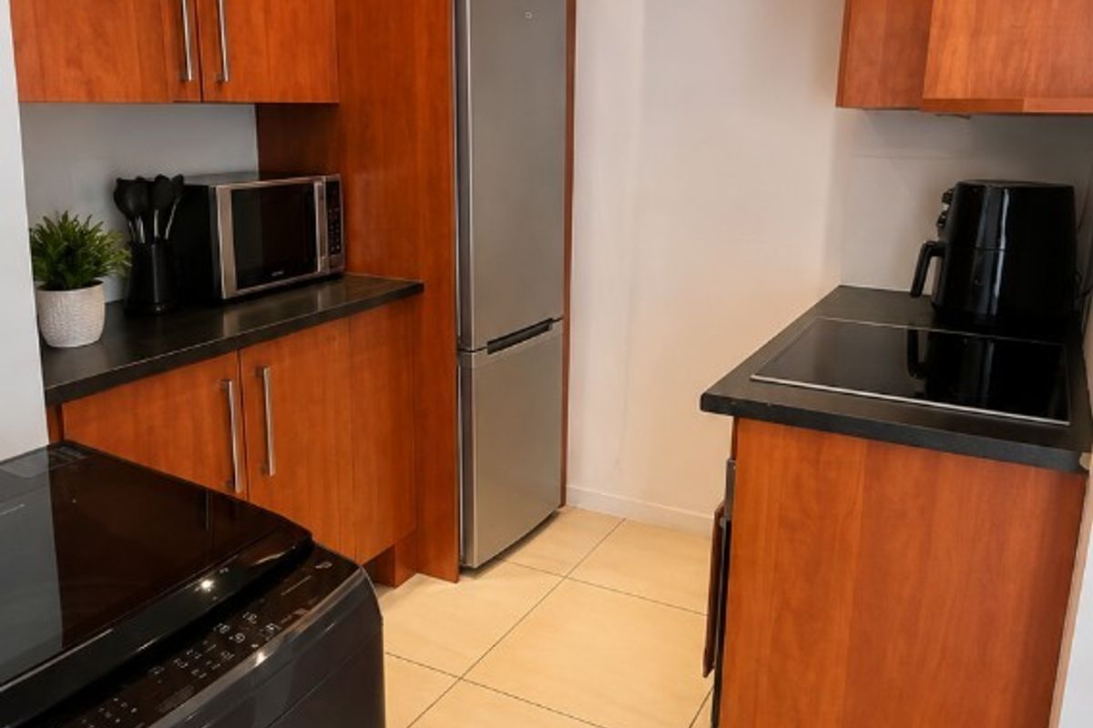 A compact kitchen featuring warm-toned wood cabinetry, dark countertops, ceramic floor tiles, and integrated appliances including an electric cooktop and extractor fan.