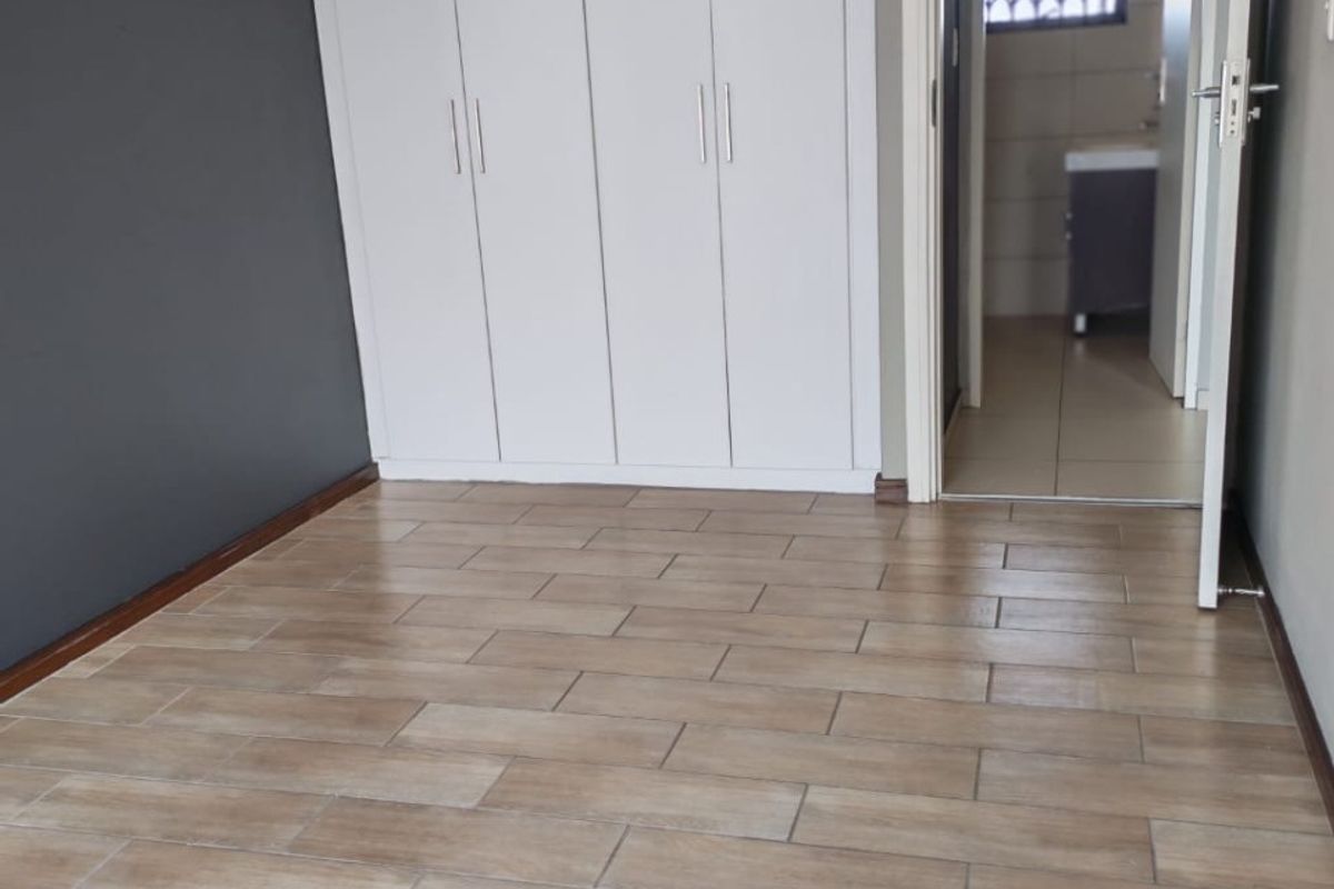 A clean, empty bedroom featuring modern wood-look floor tiles, built-in white cupboards, and an adjoining bathroom entrance.