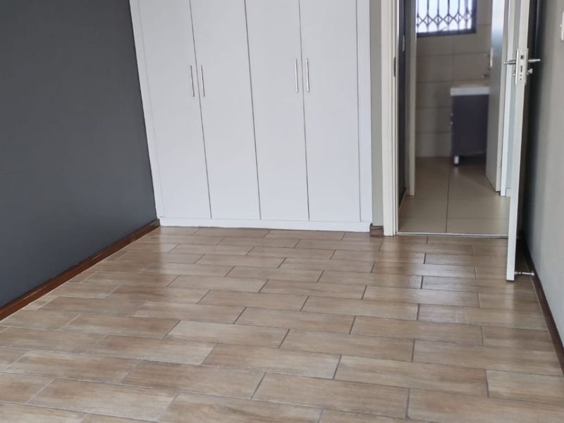 A clean, empty bedroom featuring modern wood-look floor tiles, built-in white cupboards, and an adjoining bathroom entrance.