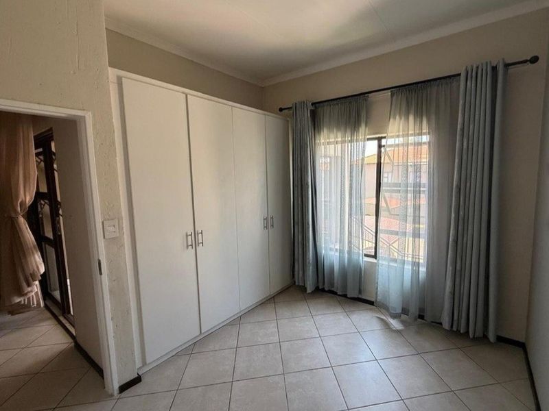 A bright, empty bedroom featuring neutral tiled flooring and ample built-in cupboard storage, with natural light entering through a window with curtain rails.