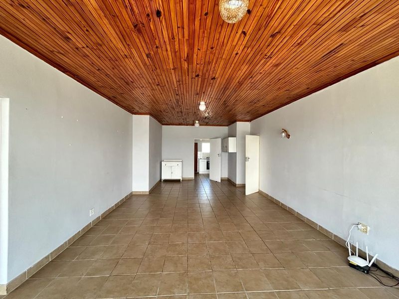 An empty living area featuring a distinctive timber tongue and groove ceiling and neutral ceramic floor tiles, leading through to the kitchen area.