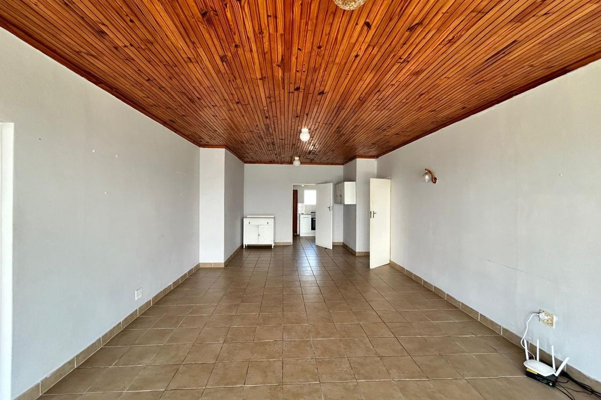 An empty living area featuring a distinctive timber tongue and groove ceiling and neutral ceramic floor tiles, leading through to the kitchen area.