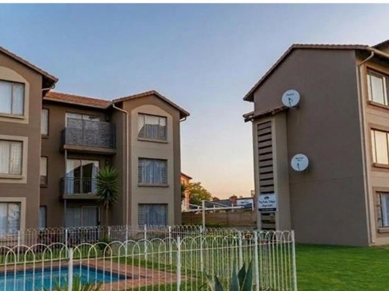 Exterior view of apartment complex buildings featuring balconies, a communal swimming pool, and landscaped grounds with perimeter fencing.