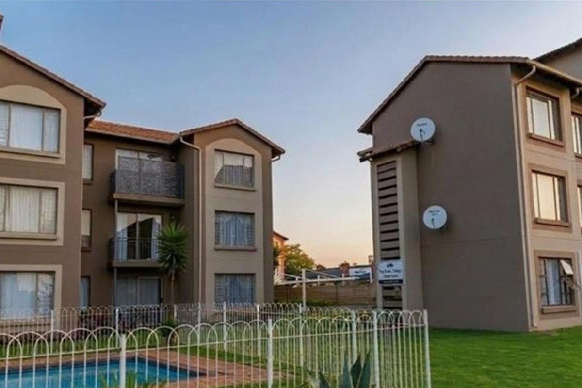 Exterior view of apartment complex buildings featuring balconies, a communal swimming pool, and landscaped grounds with perimeter fencing.