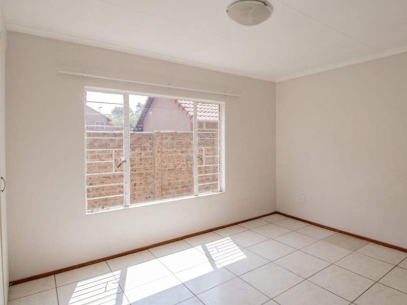 A bright, empty bedroom featuring neutral tiled flooring, a built-in cupboard, and security bars on the window.