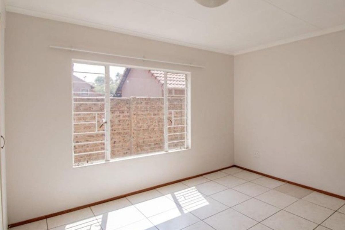 A bright, empty bedroom featuring neutral tiled flooring, a built-in cupboard, and security bars on the window.