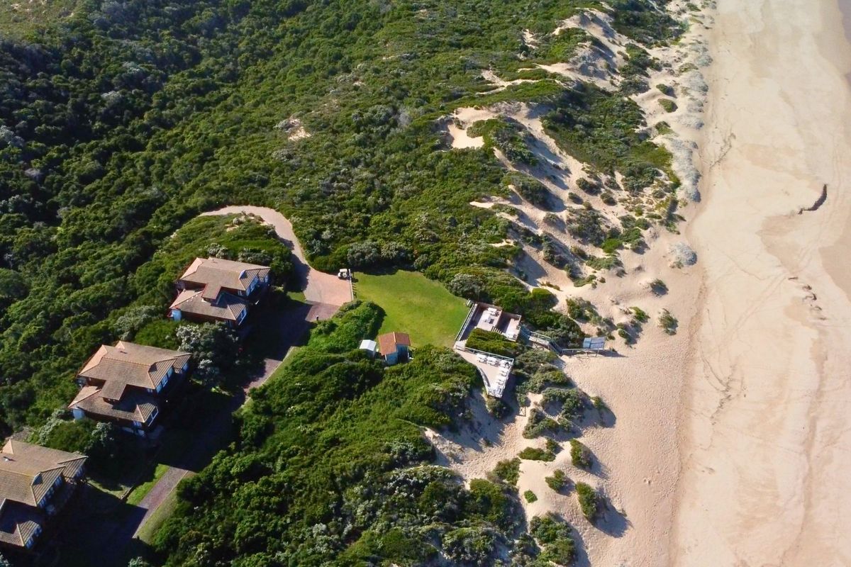 Aerial view of a coastal property complex situated directly along the beach, featuring multiple residential structures, landscaped grounds, and direct access to the coastline.