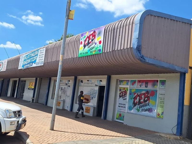 Front elevation of a commercial strip mall featuring multiple storefronts with roller shutter doors, signage, and a paved pedestrian walkway.