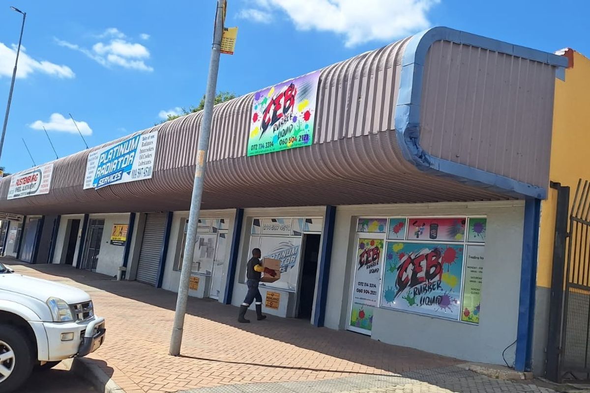 Front elevation of a commercial strip mall featuring multiple storefronts with roller shutter doors, signage, and a paved pedestrian walkway.