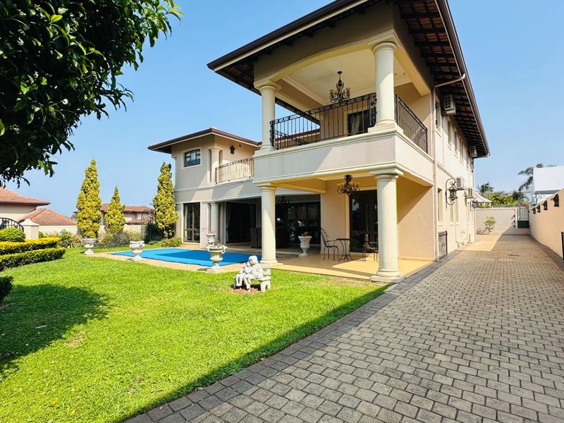 Rear view of a two-story house featuring a covered patio, upper balcony, swimming pool, and a well-maintained paved driveway and garden.