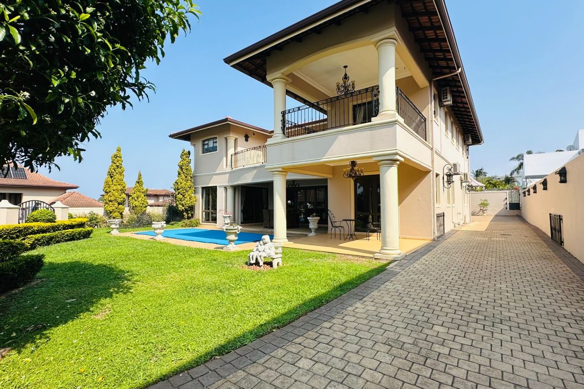 Rear view of a two-story house featuring a covered patio, upper balcony, swimming pool, and a well-maintained paved driveway and garden.