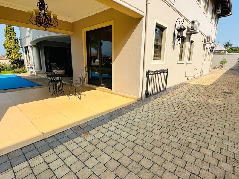 A spacious covered patio area featuring tiled flooring, overlooking a swimming pool, with adjacent paved driveway and exterior wall-mounted lighting.