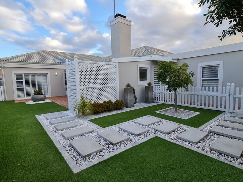 A well-maintained rear garden featuring artificial turf, stone paver pathways, white picket fencing, and a privacy screen, complemented by a clean exterior facade.