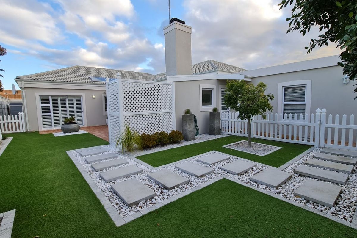 A well-maintained rear garden featuring artificial turf, stone paver pathways, white picket fencing, and a privacy screen, complemented by a clean exterior facade.