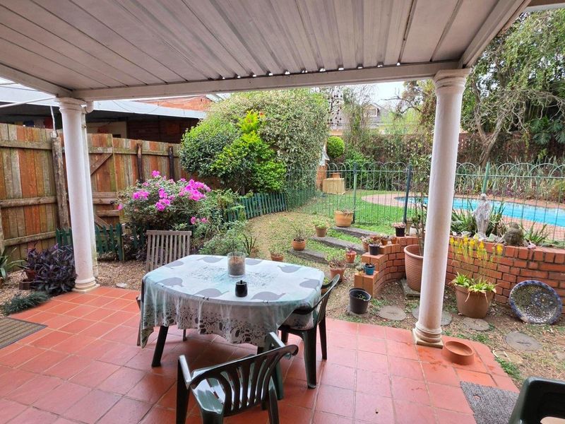 A covered outdoor patio area featuring tiled flooring, overlooking a garden with a brick retaining wall and a swimming pool.