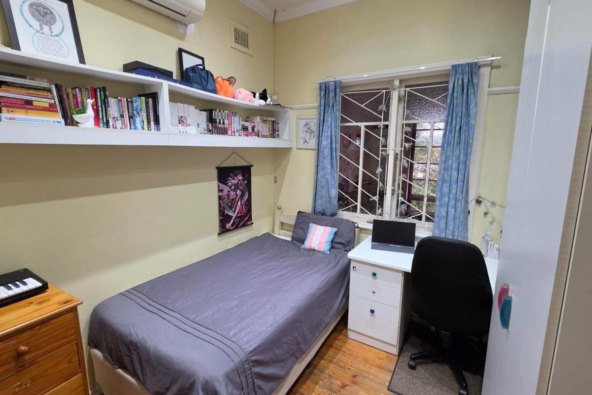 A functional bedroom featuring timber flooring, built-in shelving, an air conditioning unit, and a window with security gate protection.