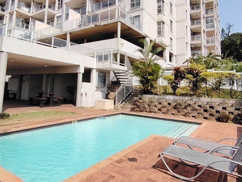 Communal swimming pool area featuring a paved deck, retaining wall, and access to the apartment building's covered patio and upper levels.