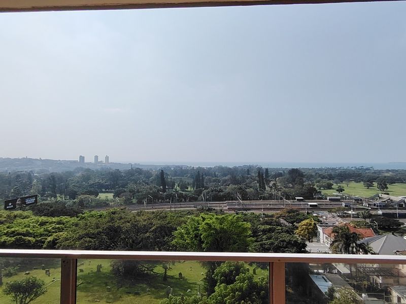 Elevated panoramic view overlooking a lush green landscape, railway infrastructure, and distant coastal horizon from a balcony.