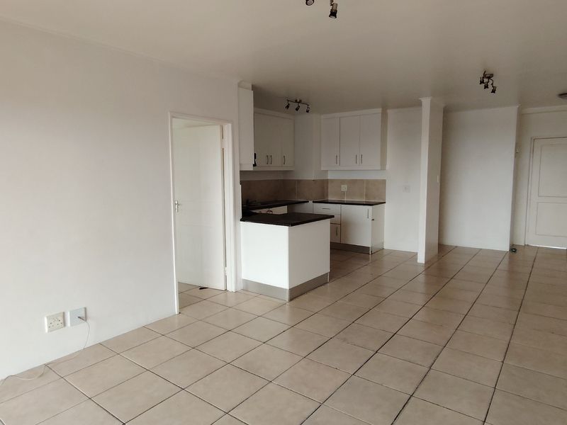 An open-plan living and kitchen area featuring neutral tiled flooring, white cabinetry, and dark countertops in a clean, vacant space.