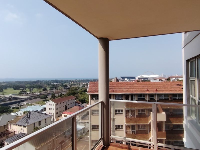 A covered balcony featuring a glass balustrade and concrete pillar, offering expansive elevated views of the surrounding neighborhood and distant coastline.
