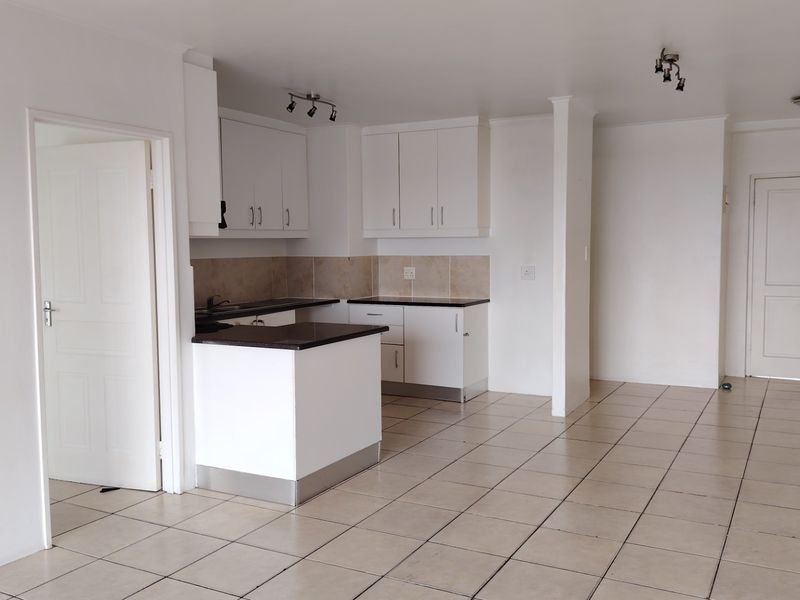 An open-plan living and kitchen area featuring neutral tiled flooring, white cabinetry, and dark laminate countertops in a clean, vacant space.