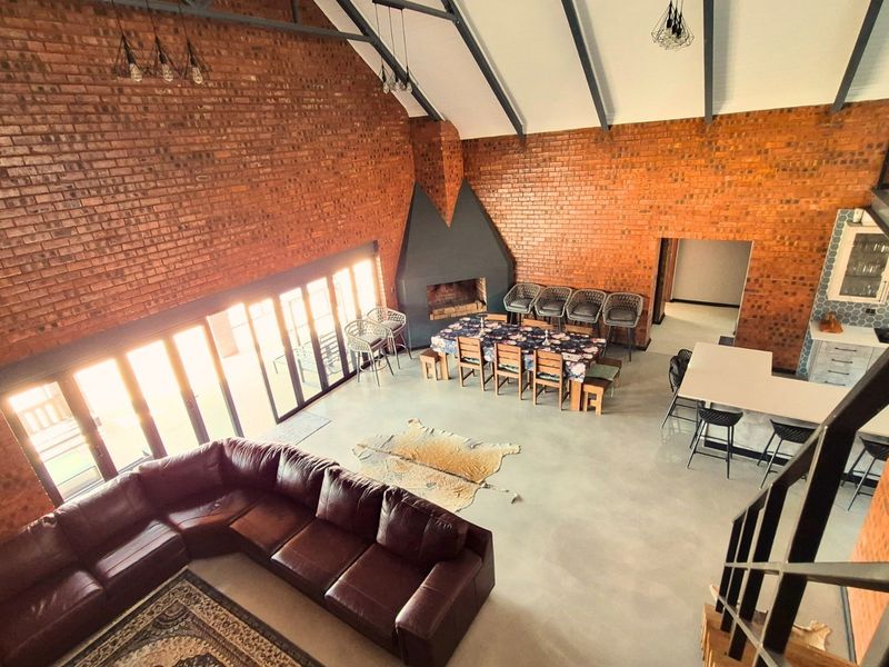 Spacious open-plan living and dining area featuring high vaulted ceilings, exposed brickwork, polished concrete floors, and a large built-in fireplace.