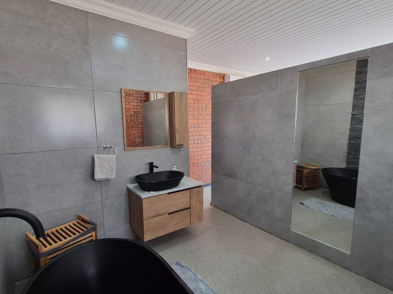 Modern bathroom featuring a sleek freestanding black bathtub, wall-mounted vanity with vessel sink, large mirror, and contemporary grey tiling with an exposed brick accent wall.