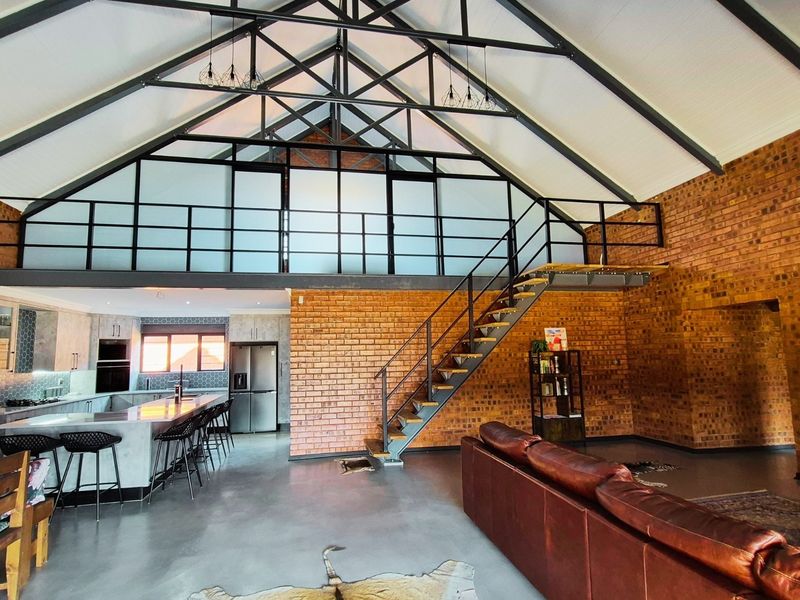 Spacious open-plan living area featuring high vaulted ceilings with exposed steel trusses, a mezzanine level, industrial-style staircase, and a modern kitchen with island.