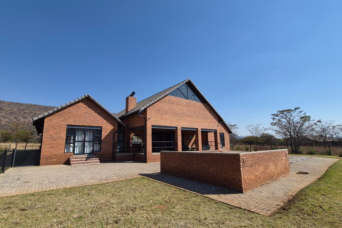Rear view of a face-brick house featuring a covered patio, paved outdoor area, and a low brick wall, set against a natural landscape.