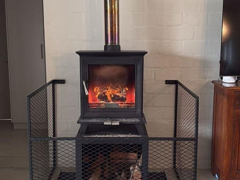 A living room featuring a freestanding wood-burning fireplace with a metal safety guard and exposed flue against a painted brick wall.