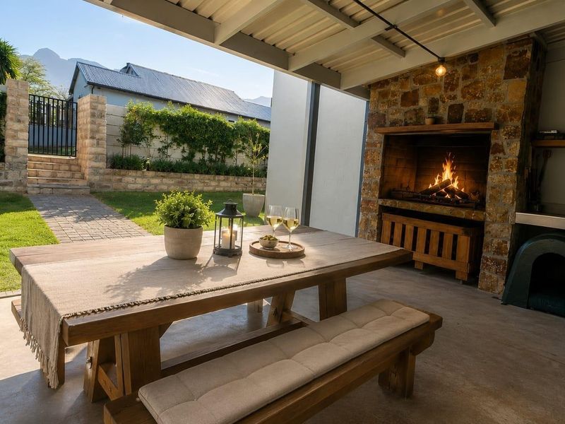 A covered outdoor patio featuring a built-in stone braai, concrete flooring, and direct access to a landscaped garden and paved walkway.