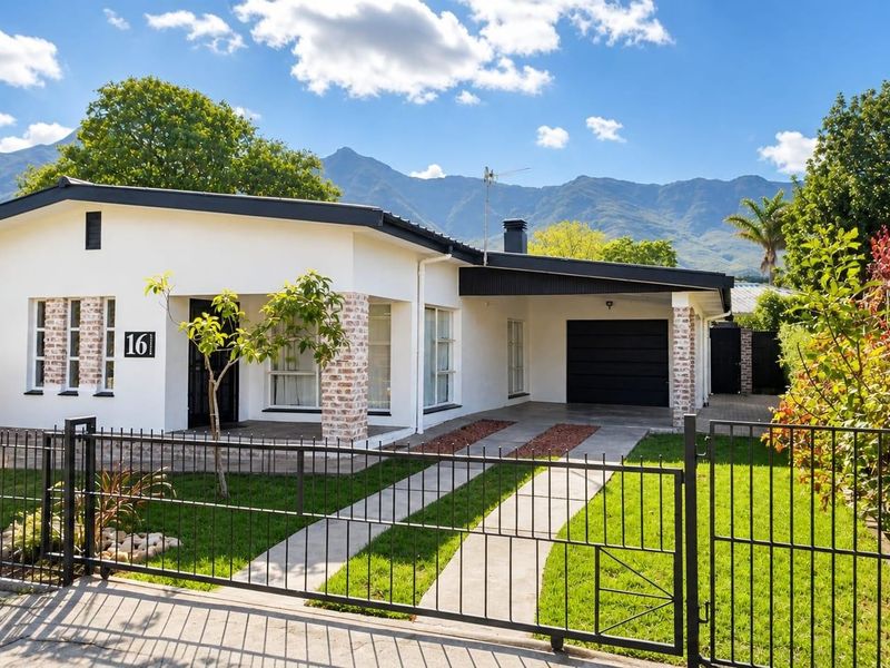 A well-maintained single-story house featuring a white facade, brick-accented pillars, a covered carport, and a neat front garden with mountain views.