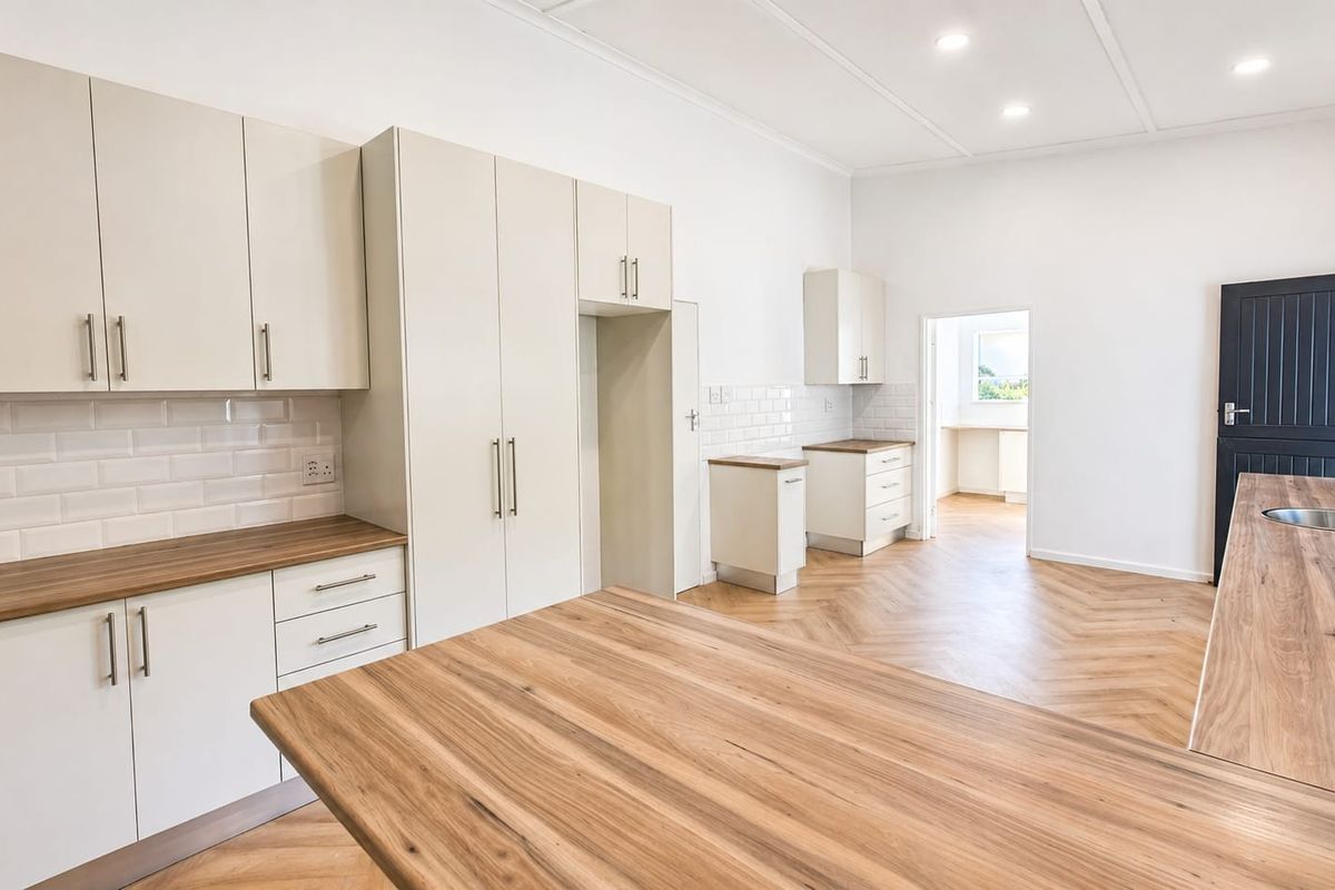 A bright, modern kitchen featuring neutral cabinetry, wood-look laminate countertops, subway tile backsplash, and herringbone flooring, with an open layout leading to an adjacent room.