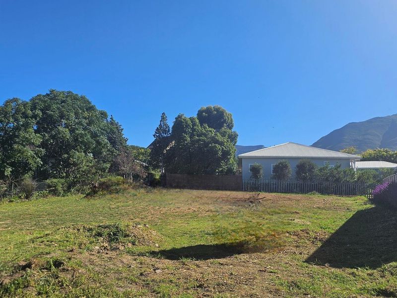 A vacant residential plot featuring established trees, perimeter fencing, and a clear view of the surrounding mountain landscape.