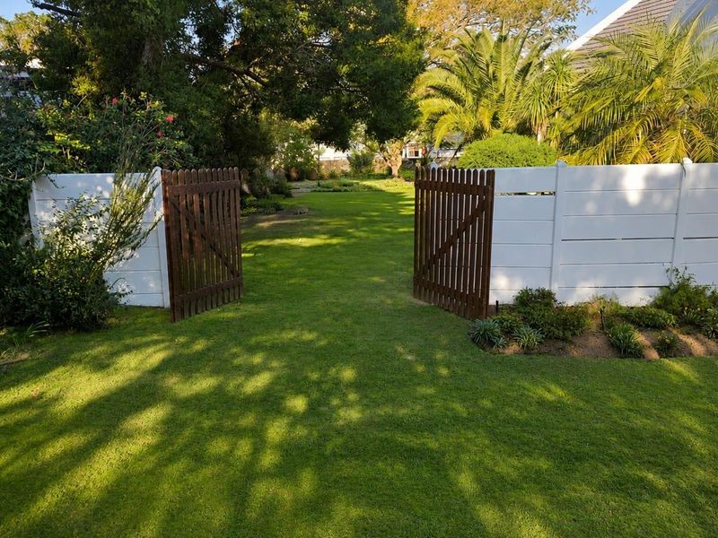 A well-maintained residential garden featuring a lush green lawn, mature trees, and a white vinyl fence with a wooden gate entrance.