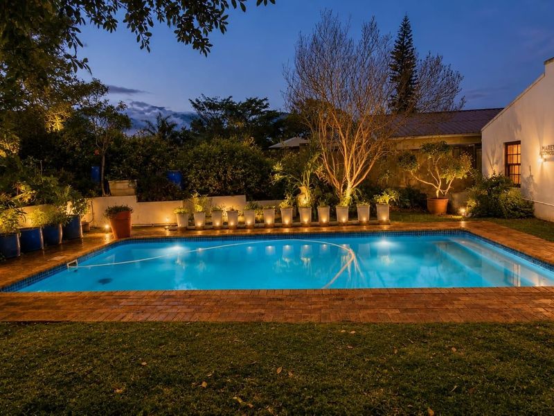 A well-maintained outdoor pool area featuring brick paving, integrated garden lighting, and a landscaped backyard setting at dusk.