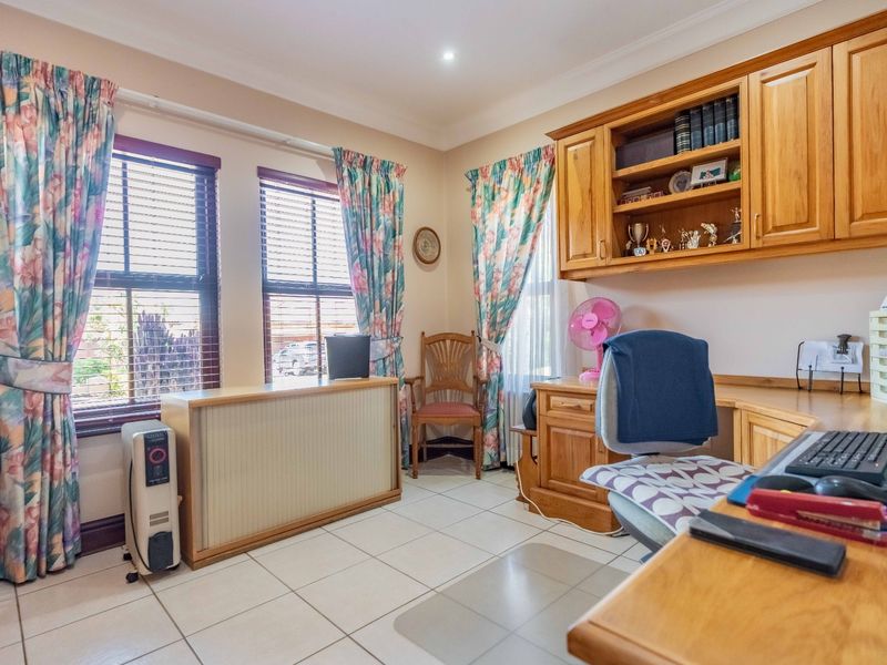A functional home office featuring built-in wooden cabinetry, a spacious desk, tiled flooring, and large windows with venetian blinds.