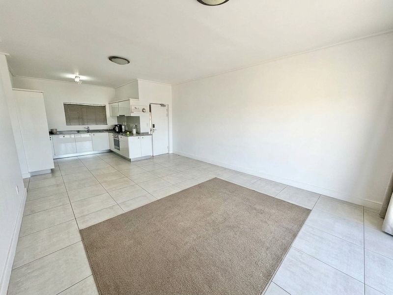 An open-plan living and kitchen area featuring neutral tiled flooring, white cabinetry, and integrated kitchen appliances in a clean, vacant space.