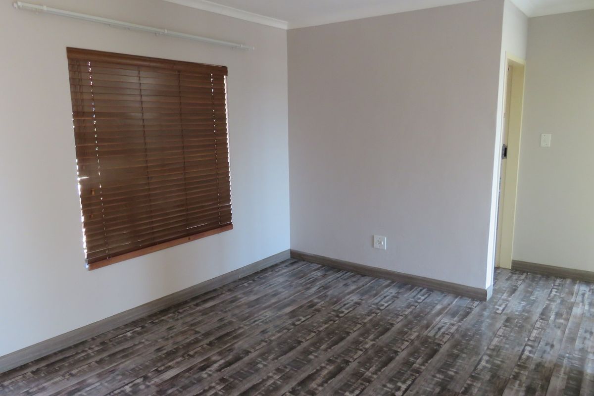 A bright, empty bedroom featuring modern laminate flooring, neutral wall tones, and fitted wooden venetian blinds.