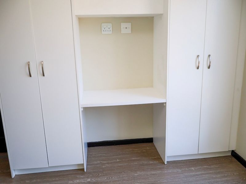 A clean bedroom featuring white built-in cupboards and a central built-in desk area with integrated electrical points and modern laminate flooring.