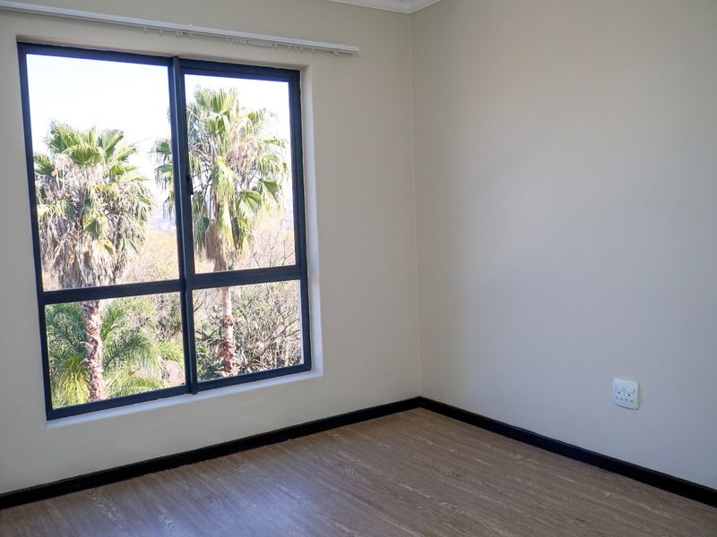 A bright, empty bedroom featuring modern laminate flooring, neutral walls, and a large window with views of palm trees.