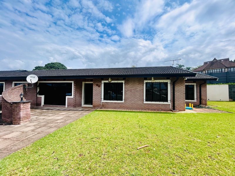 Rear view of a single-story brick house featuring a tiled roof, paved patio area, and a well-maintained lawn.