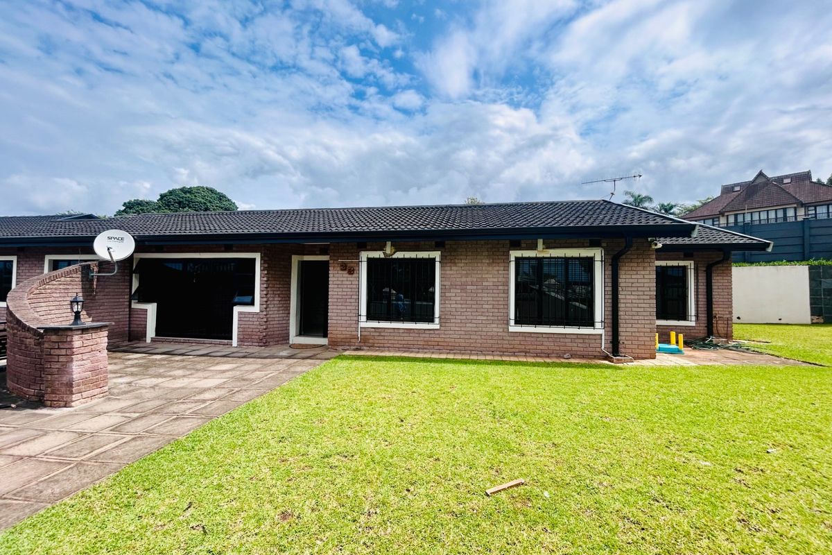 Rear view of a single-story brick house featuring a tiled roof, paved patio area, and a well-maintained lawn.